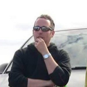 A man wearing dark sunglasses rests his hand on his chin, standing in front of a car under a cloudy sky, contemplating the upcoming drone show.