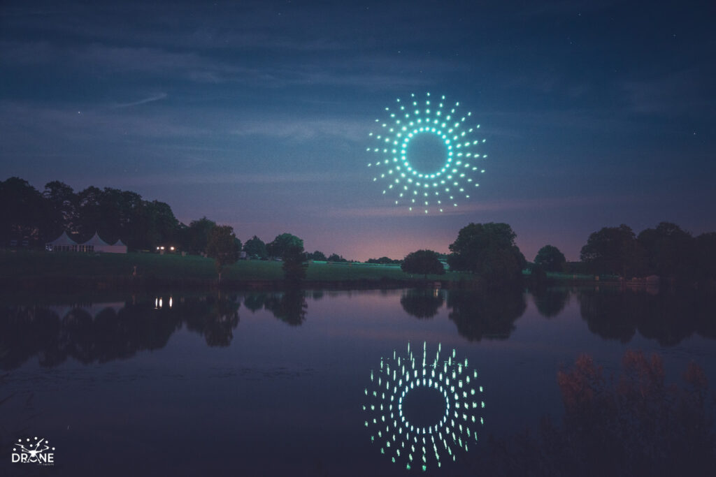 A mesmerizing drone light show fills the night sky, with drones casting green lights and forming a circle pattern above a calm lake, their reflections shimmering on the water.