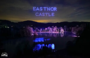 A nighttime drone light show spells "EASTNOR CASTLE" in blue lights over a dark, calm lake surrounded by trees.