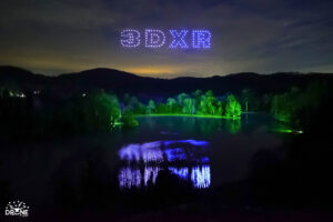 Night sky over a lake with an illuminated "3DXR" drone light show, trees reflecting on water, and forested hills in the background.