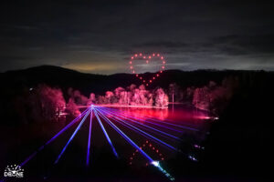 A nighttime scene with laser lights over a lake, glowing trees, and a mesmerizing drone light show where drones form a heart shape in the sky.