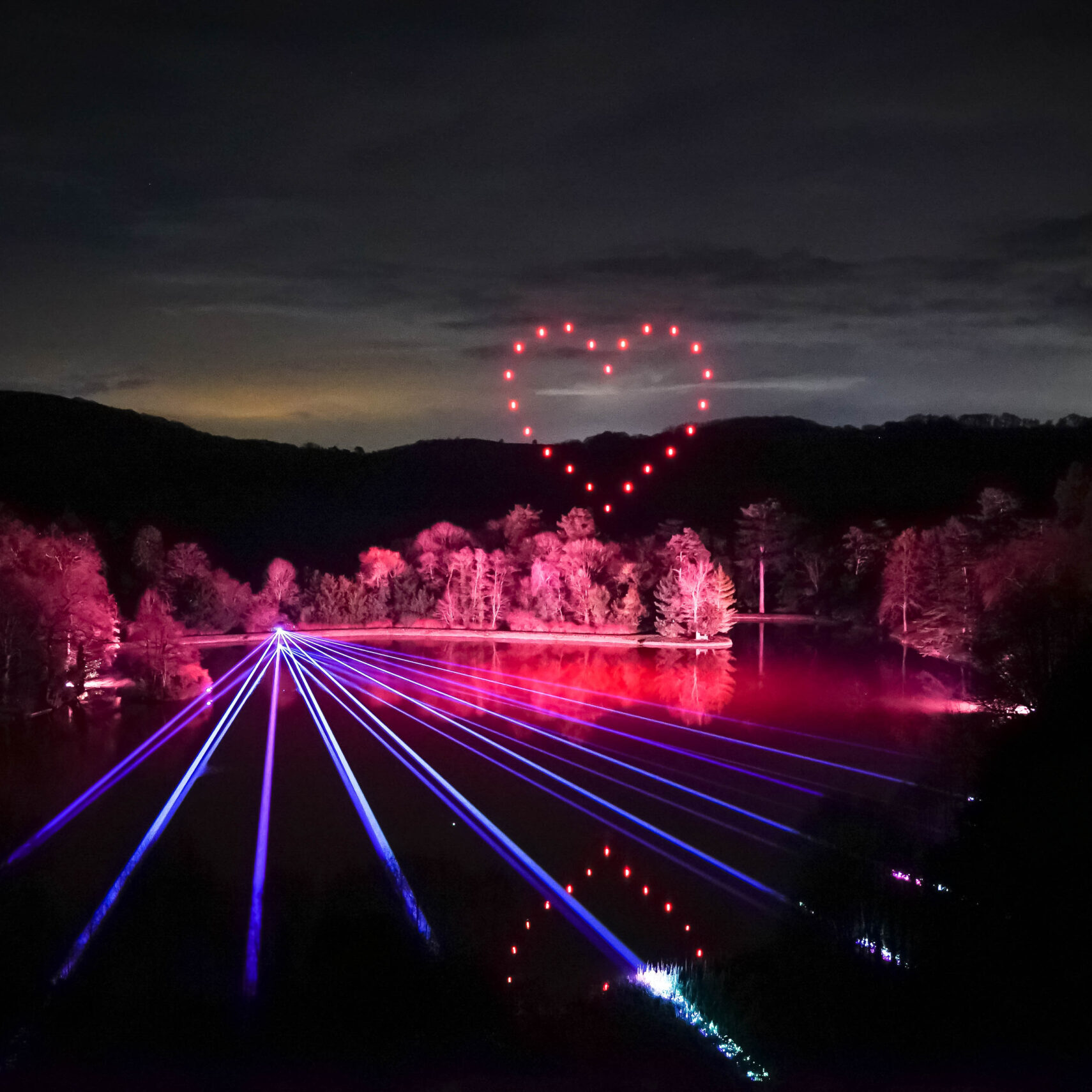 A nighttime light show with lasers over a lake, forming a red heart shape in the sky.