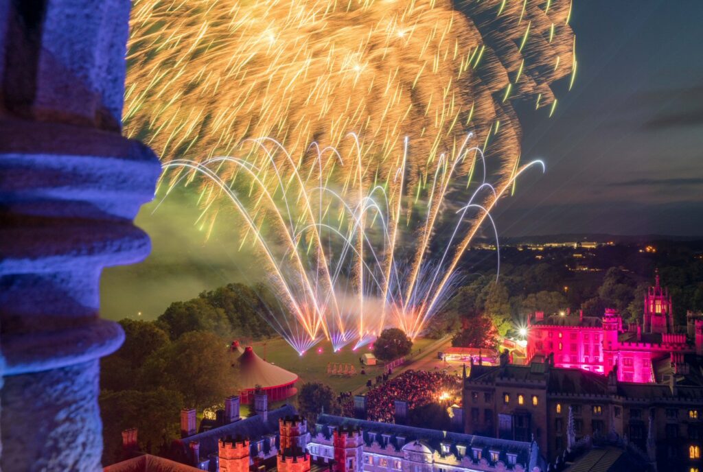 Fireworks display lighting up the night sky over a large, illuminated castle and its surrounding grounds, enhanced by a mesmerizing drone light show.