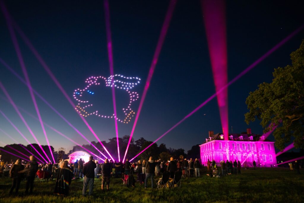 A nighttime drone light show features lights forming a rabbit shape, with vibrant purple beams projecting from a building.