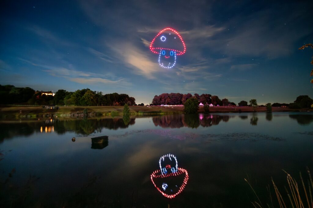 A nighttime sky features drone lights forming mushroom shapes, reflected in a calm lake below, surrounded by trees.