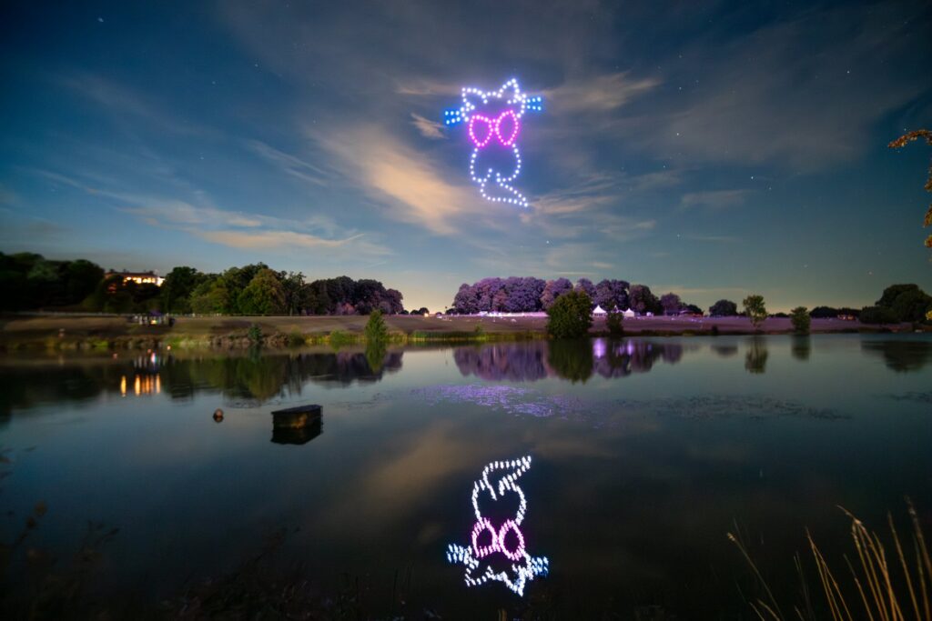 A night sky with a formation of drones creating a face with glasses, reflected in a calm lake below.