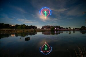 A heart-shaped drone light display with colorful patterns is reflected in a calm lake under a night sky.