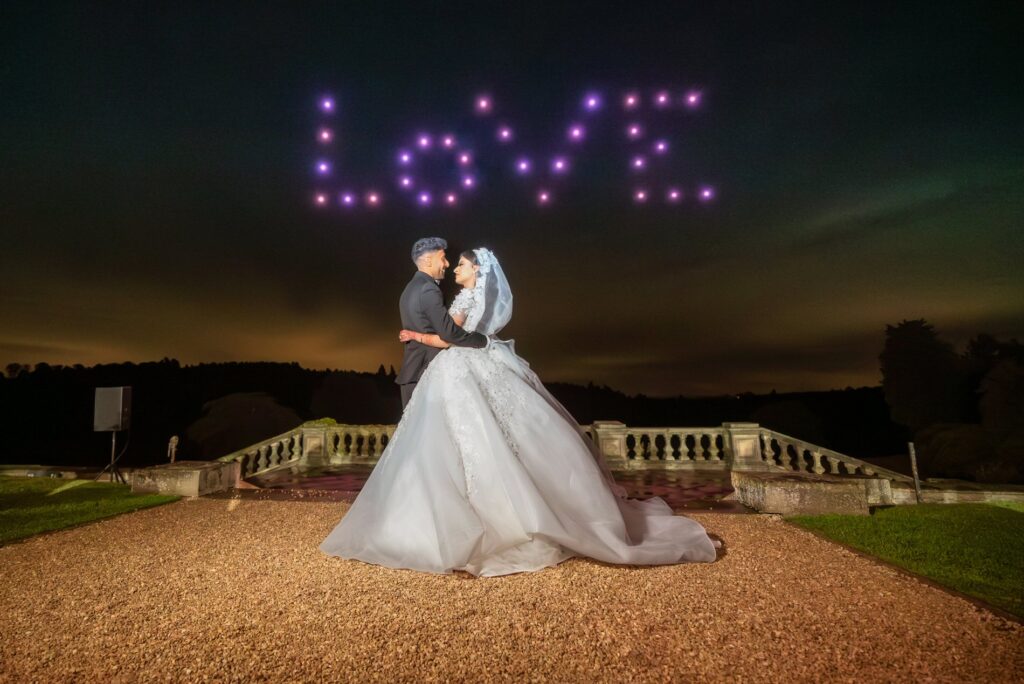 A bride and groom embrace outside at night with "LOVE" written in the sky using purple lights.