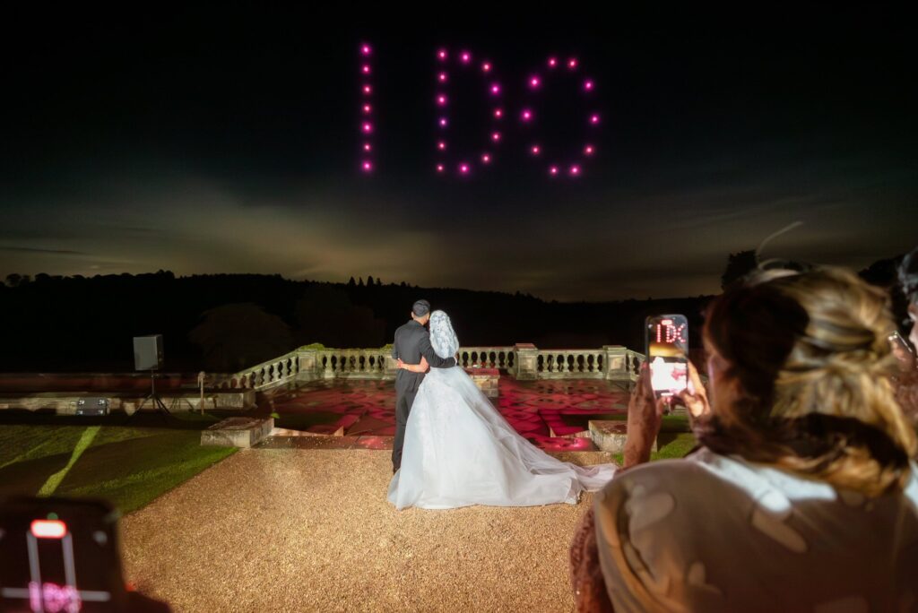 A bride and groom embrace as "I DO" is written in pink lights in the night sky, with onlookers capturing the moment.