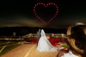 A couple in wedding attire watches a heart-shaped light display in the night sky.