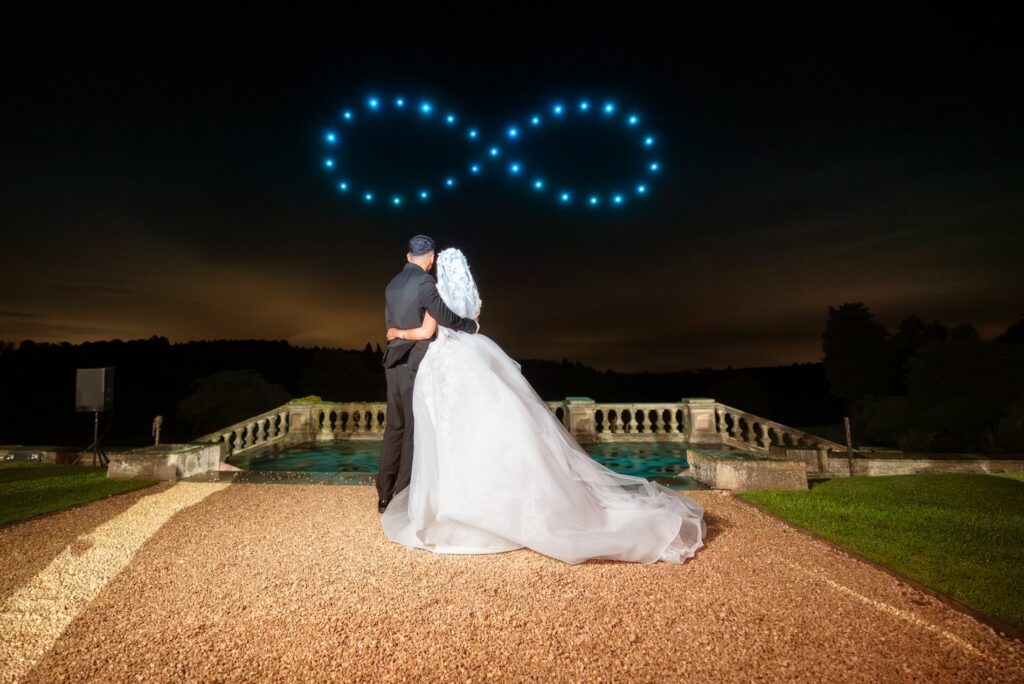 A bride and groom watch drones form an infinity symbol in the night sky during their outdoor wedding.