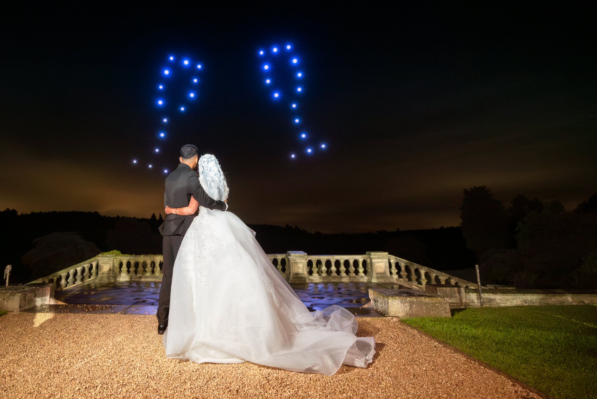 A couple in wedding attire embrace while looking at a night sky illuminated with two heart-shaped patterns made of lights.