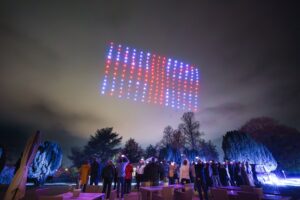 A group of people watch a colorful drone light display in the night sky.