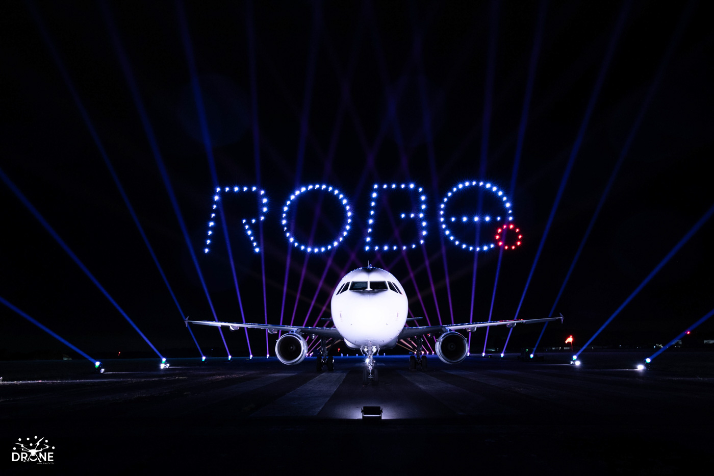 Airplane on runway with drone lights above spelling "ROBE," surrounded by colorful beams in the night sky.