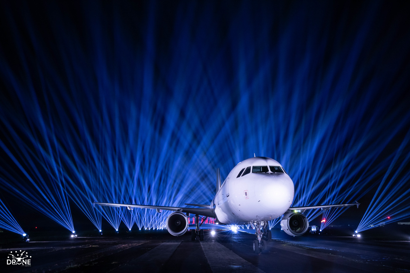 Airplane on runway lit by dramatic blue lights, with beams radiating in the night sky.