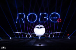 Airplane on runway with illuminated "ROBE" in drone lights above, set against a dark night sky.