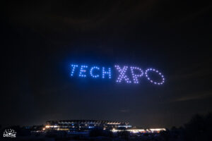 Drones form "TECH XPO" in the night sky over a stadium.