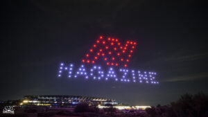 Drones form "MAGAZINE" with lights in the night sky above a stadium.