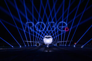 Airplane on runway with blue lights and "RODE" spelled above with illuminated drones at night.