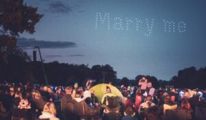 Crowd watching a nighttime sky proposal spelled by drones saying "Marry me" above a park.