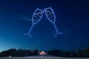 Drones light up the night sky, forming two clinking champagne glasses above a large building.