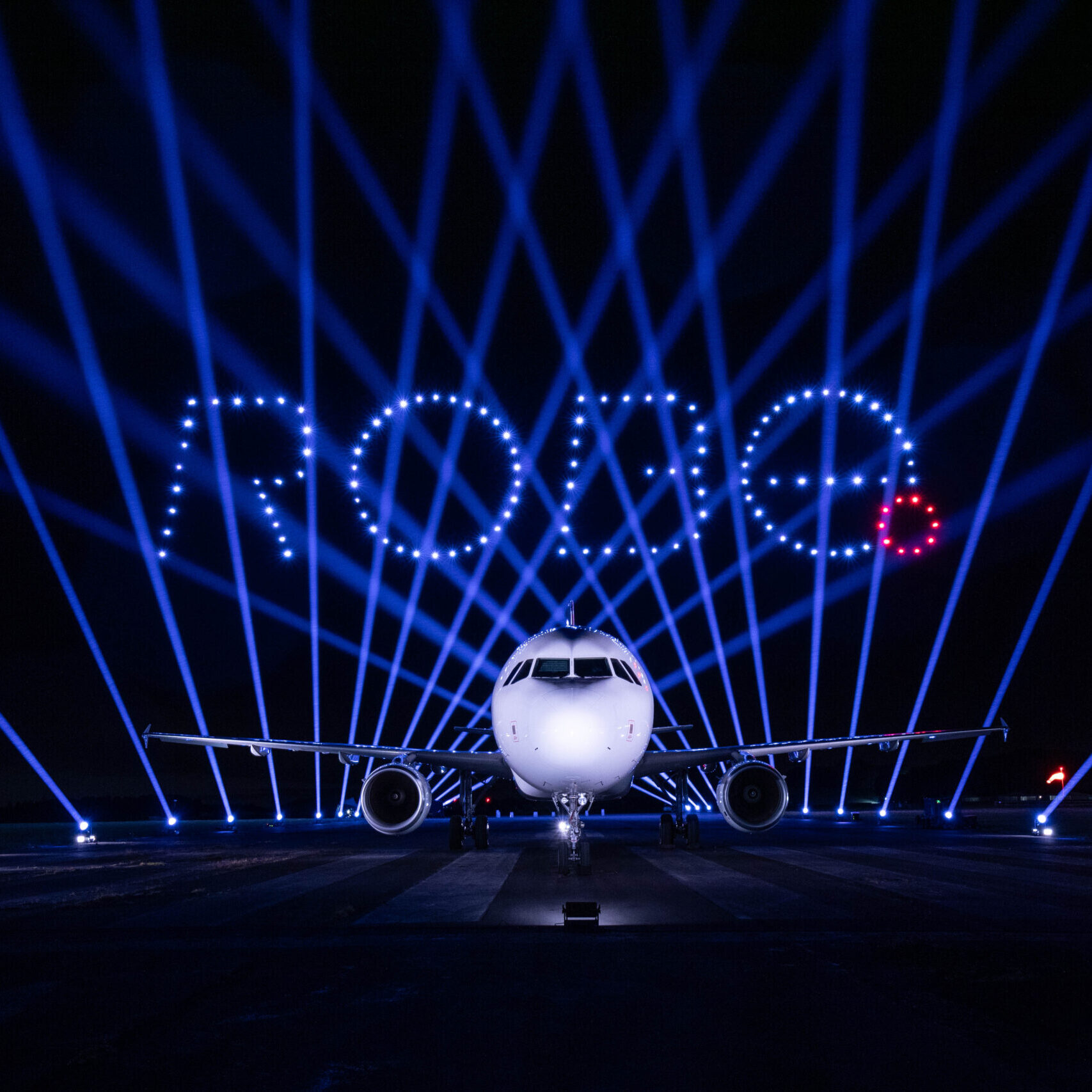 A plane on a runway with blue and red lights forming the word "ROUGE" above it at night.