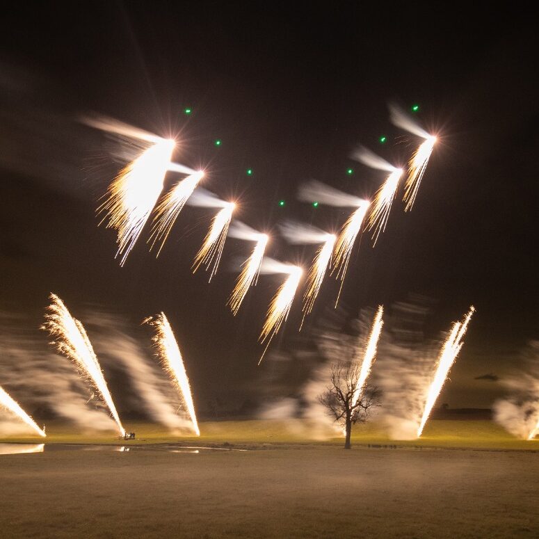 Fireworks curve in the night sky over a field with a lone tree, while a Drone Swarm System adds an extra layer of dramatic light display.