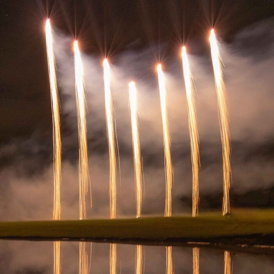 Six vertical fireworks reflecting over calm water against a dark night sky.